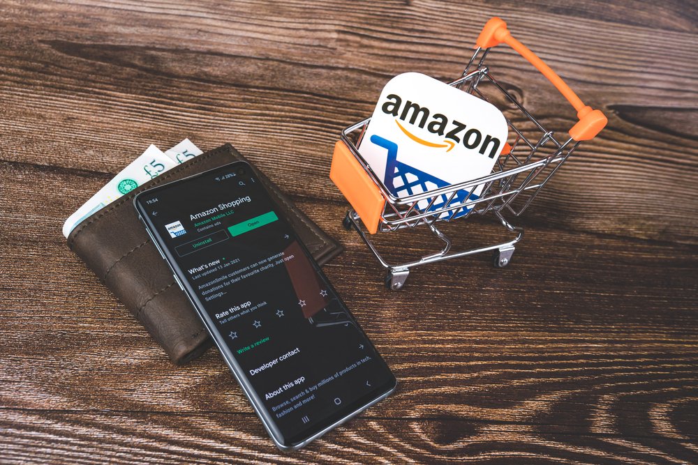 Mini shopping trolley with Amazon logo and Amazon app on Google Play on smartphone and wallet with pounds currency.